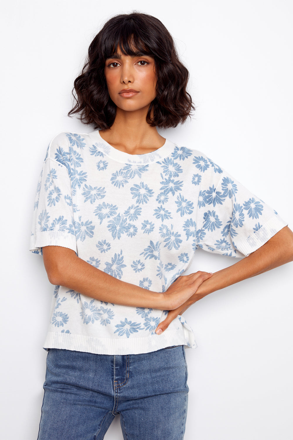 Woman wearing a white sweater with blue floral pattern and blue jeans on a white background.