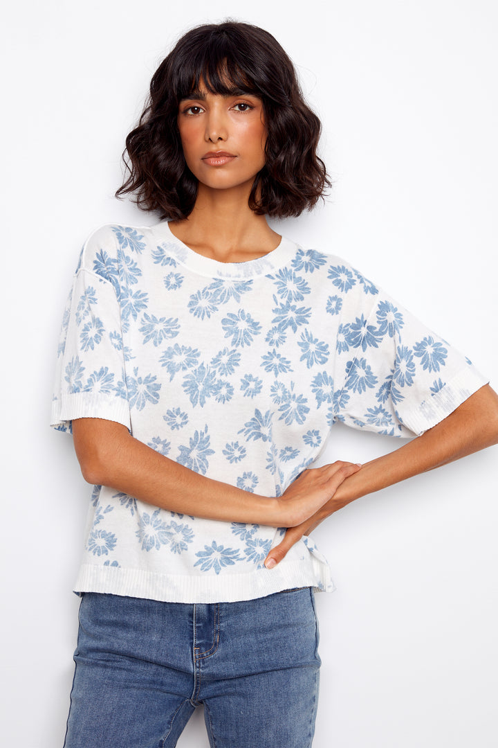 Woman wearing a white sweater with blue floral pattern and blue jeans on a white background.