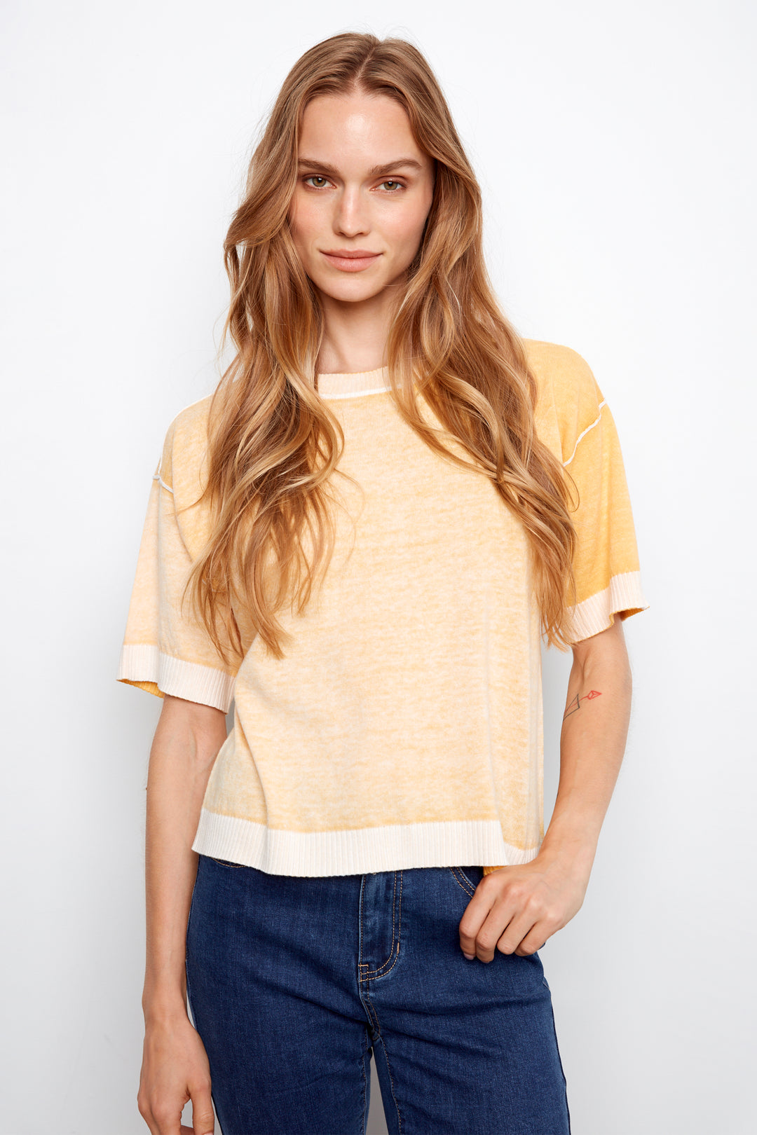Woman wearing a yellow and white top with blue jeans on a plain background