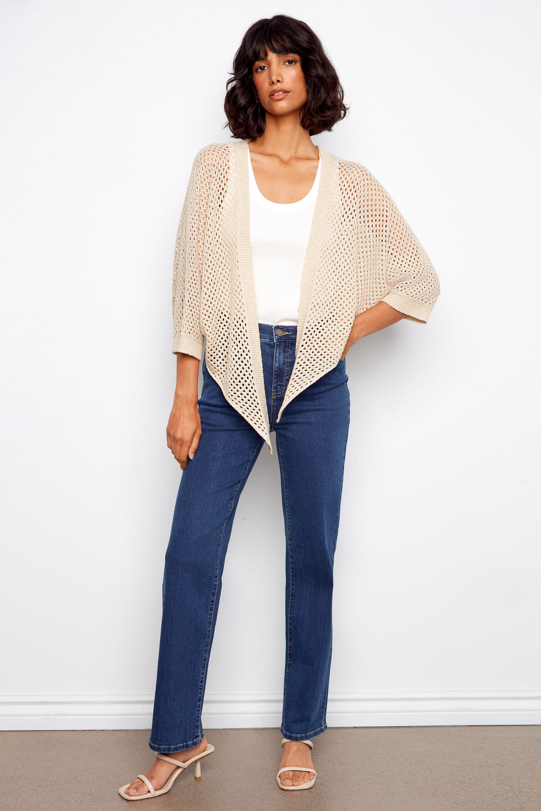 Woman wearing a beige knitted cardigan, white top, and blue jeans against a white background