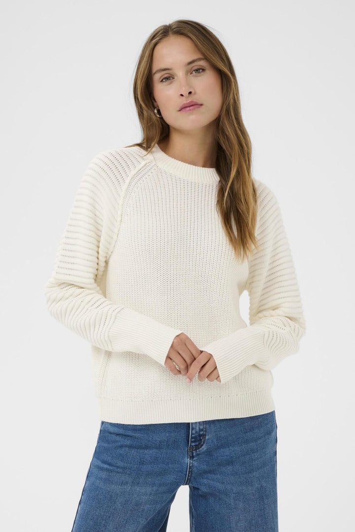 Woman wearing a cream ribbed design sweater and blue jeans against a white background