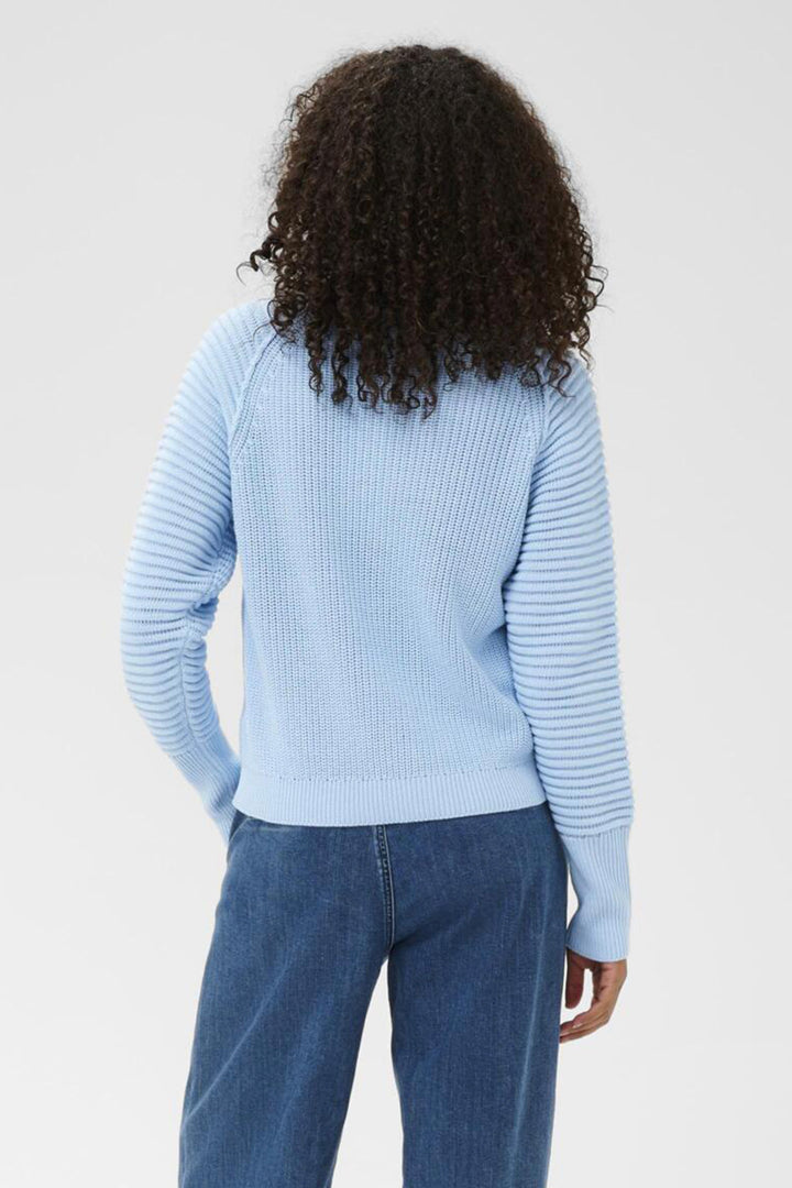 Person wearing a light blue ribbed sweater and blue jeans on a plain background