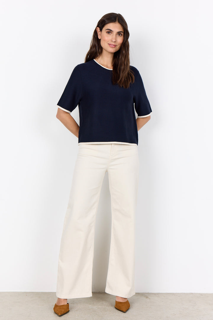 Woman wearing a navy top and white pants against a white background