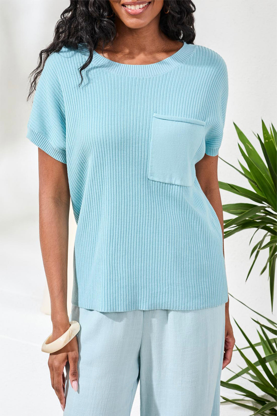 Woman wearing a light blue ribbed sweater top with a pocket, standing against a white background with green plants.