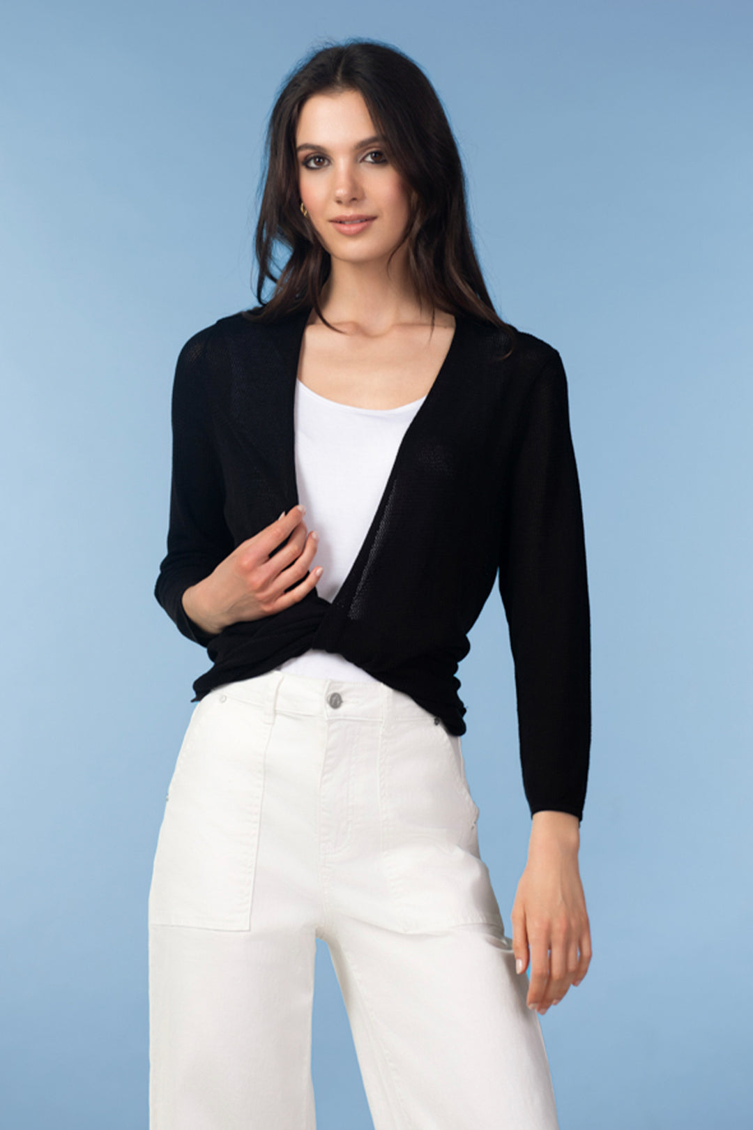 Woman wearing a black cover up cardigan over a white top and pants on a light blue background