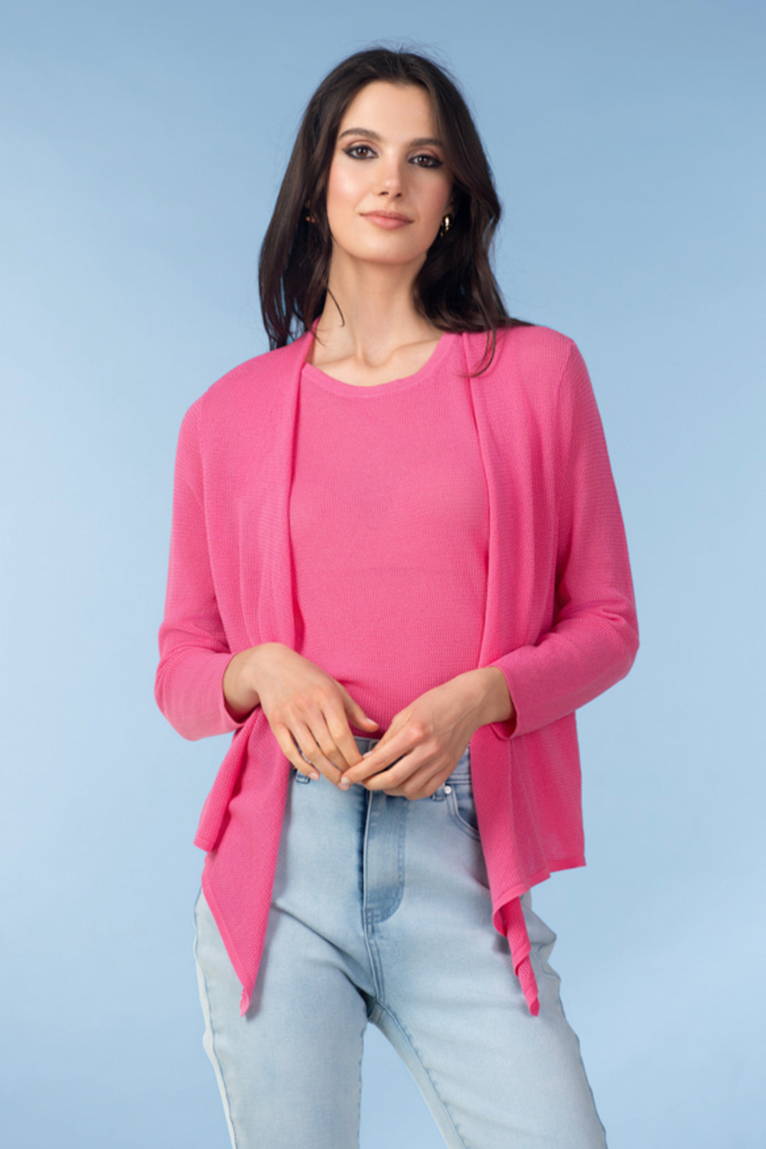 Woman wearing a pink cover up cardigan over a pink top and light blue jeans on a light blue background