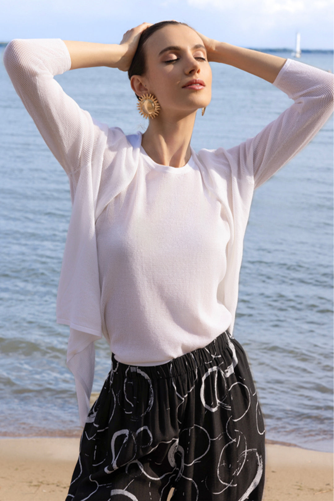 Woman standing on a beach with ocean view, wearing a white cover up cardigan and black patterned skirt.