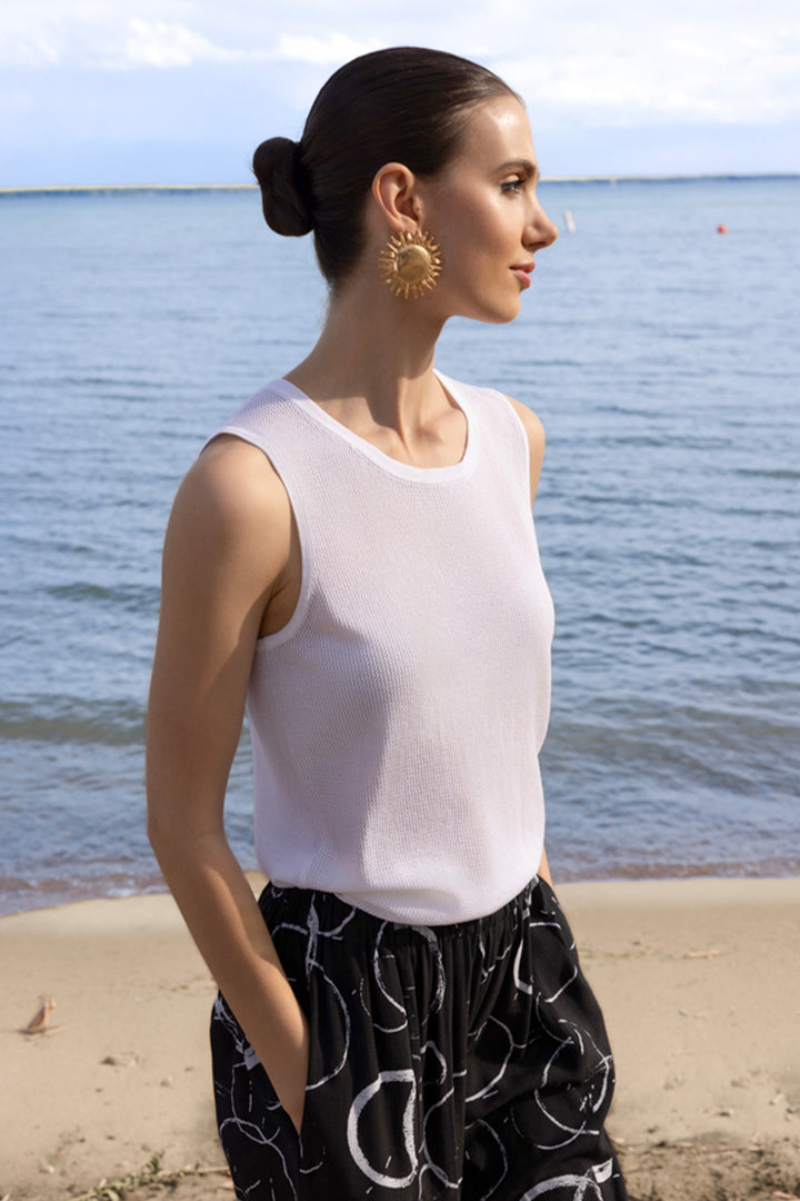 Woman standing on a beach wearing a white sleeveless top and black pants with a visible brand logo.