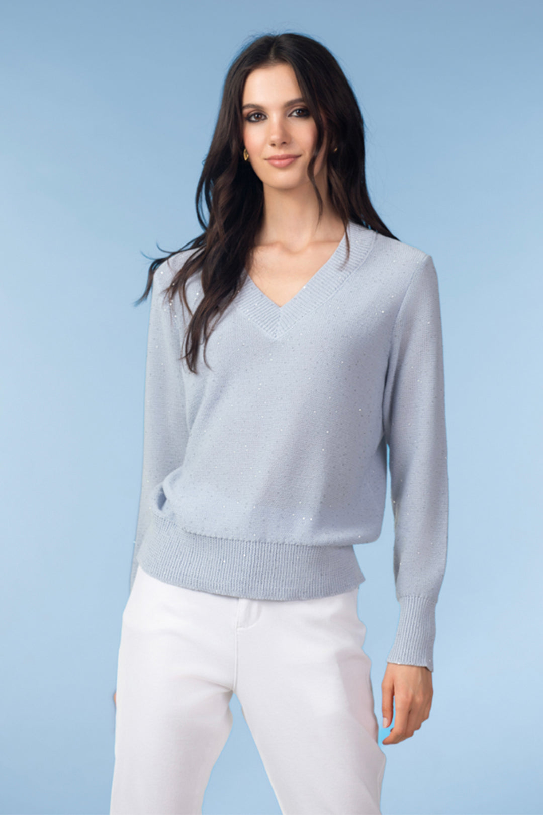 Woman wearing a light gray sweater with sequins and white pants against a light blue background