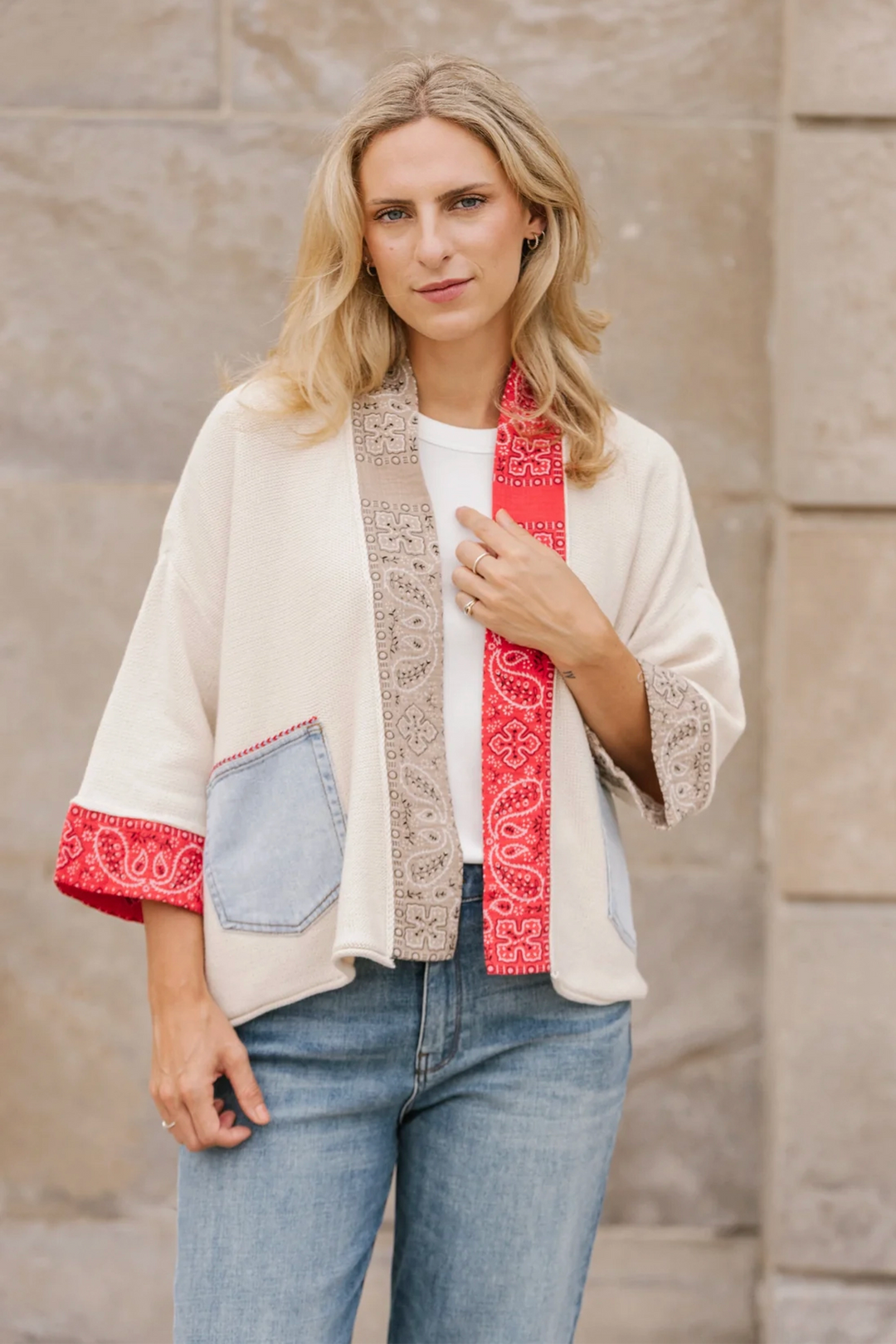 Woman wearing a cream open cardigan  with red and blue accents against a stone wall.