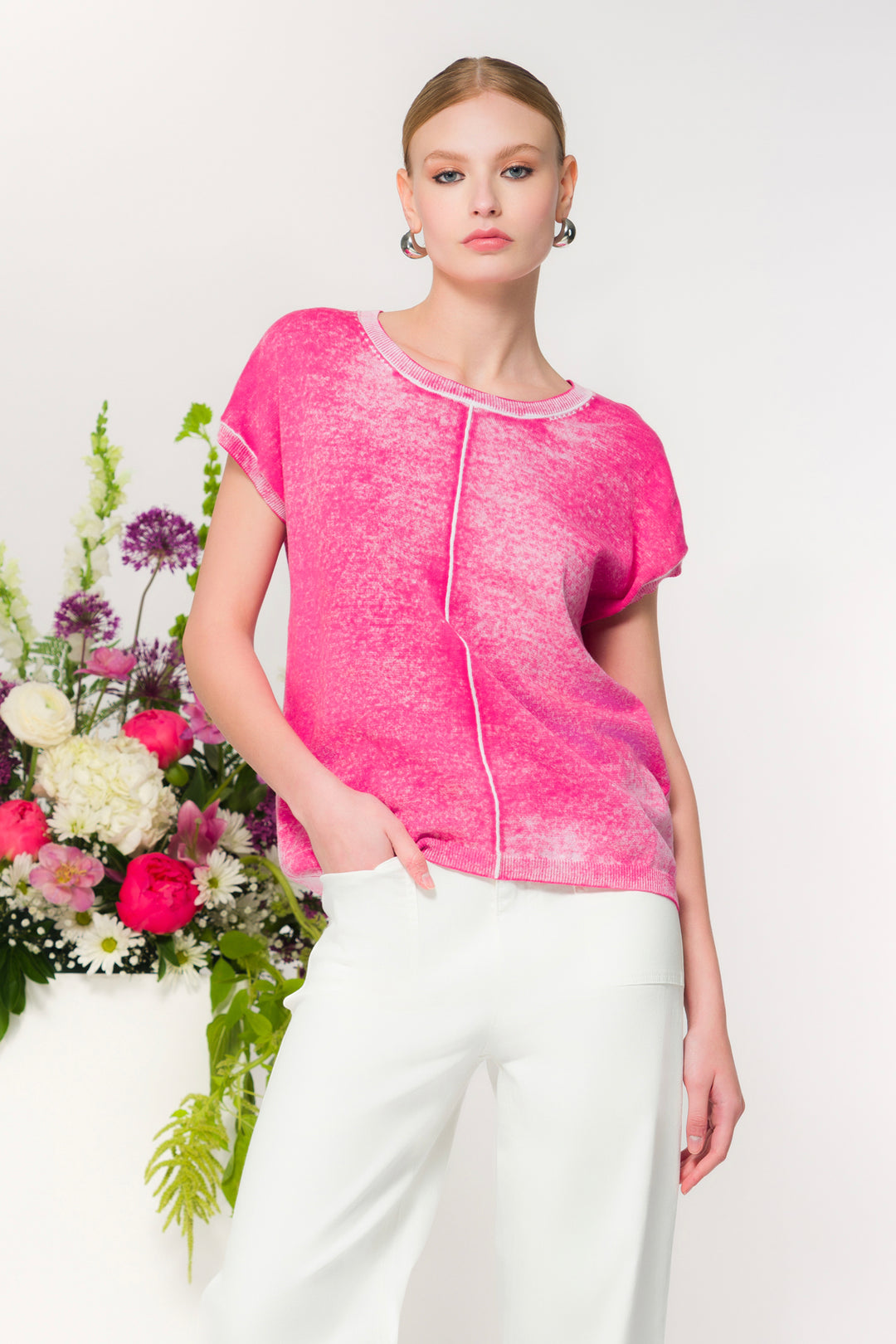 Woman wearing a pink sweater top with white pants standing next to a floral arrangement on a white background