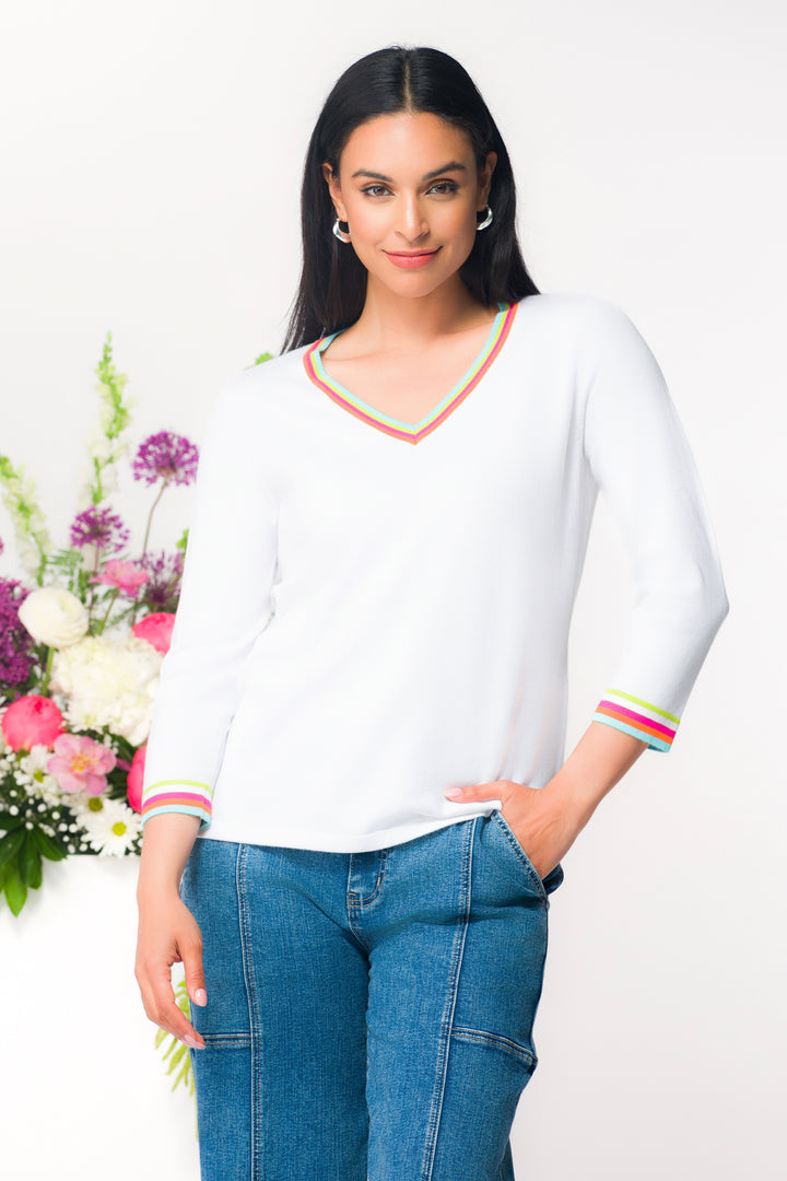 Woman wearing a white sweater top with colorful trim and blue jeans, standing next to a floral arrangement on a white background.