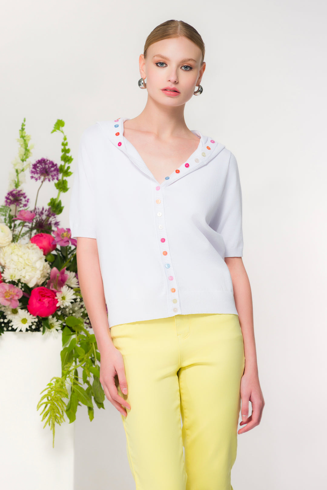 Woman wearing a button cardigan with hood white with colorful buttons and yellow pants, standing next to a floral arrangement