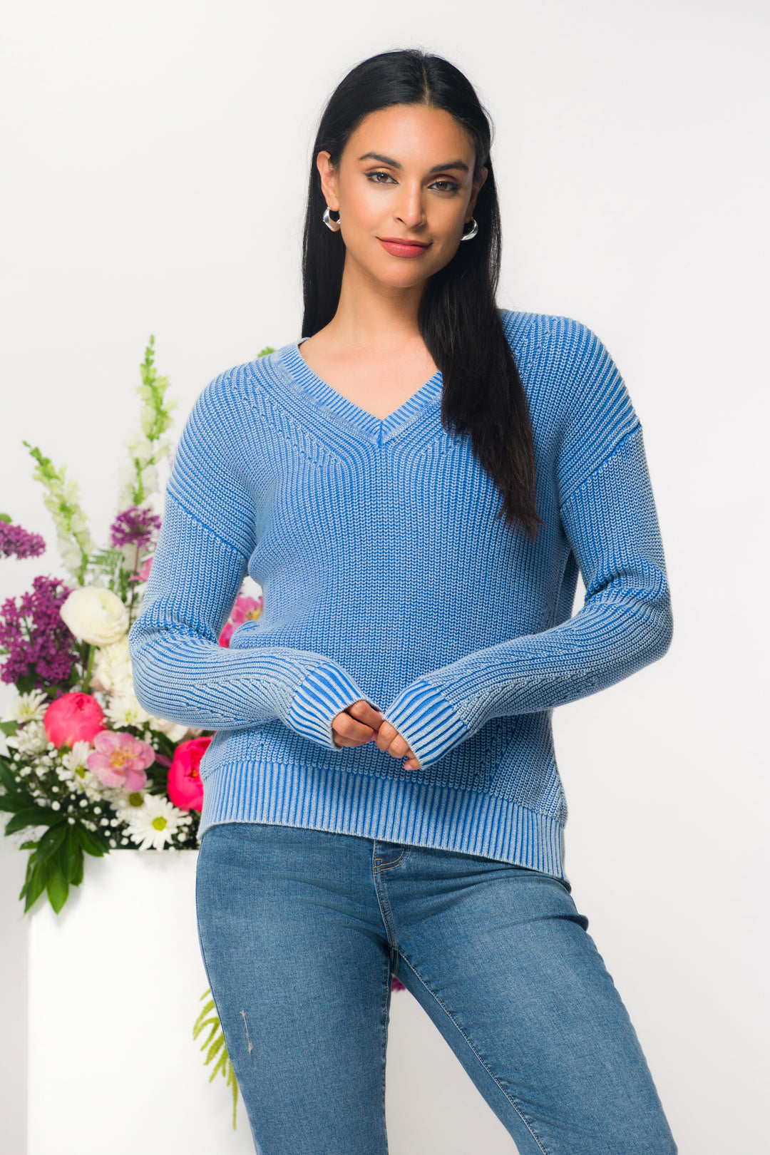Woman wearing a blue sweater and jeans standing next to a floral arrangement on a white background
