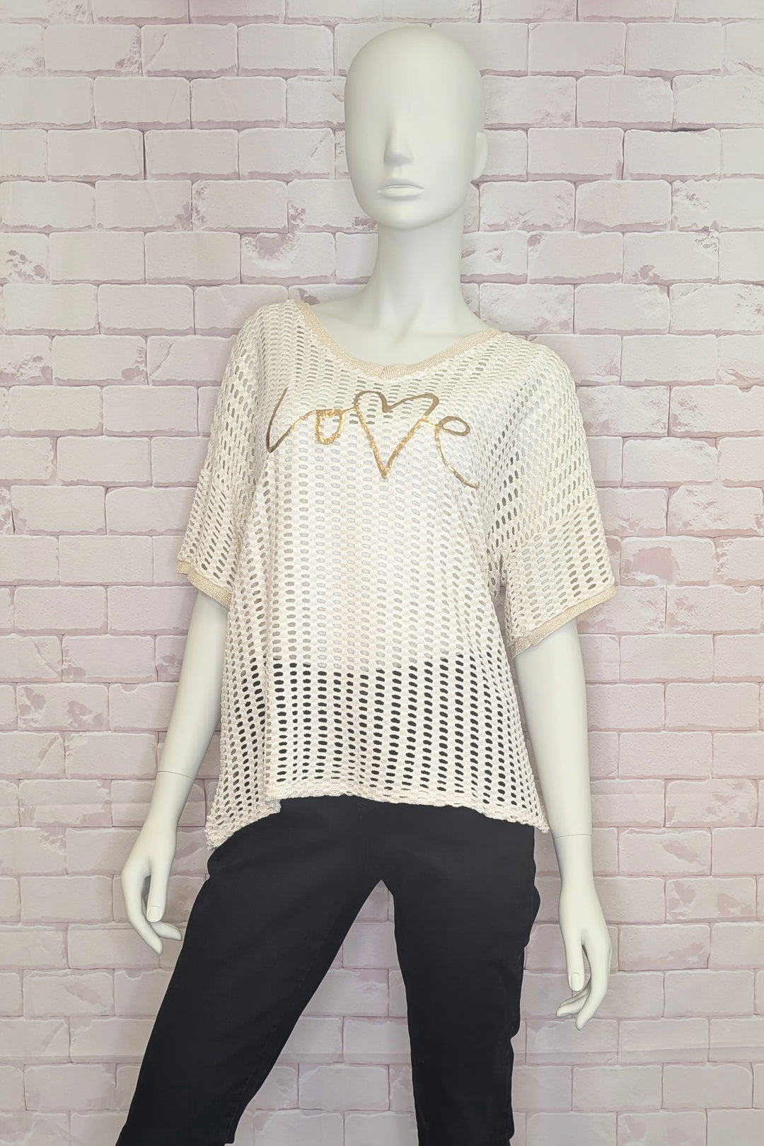 Mannequin wearing a beige crochet sweater top with 'LOVE' design against a brick wall.