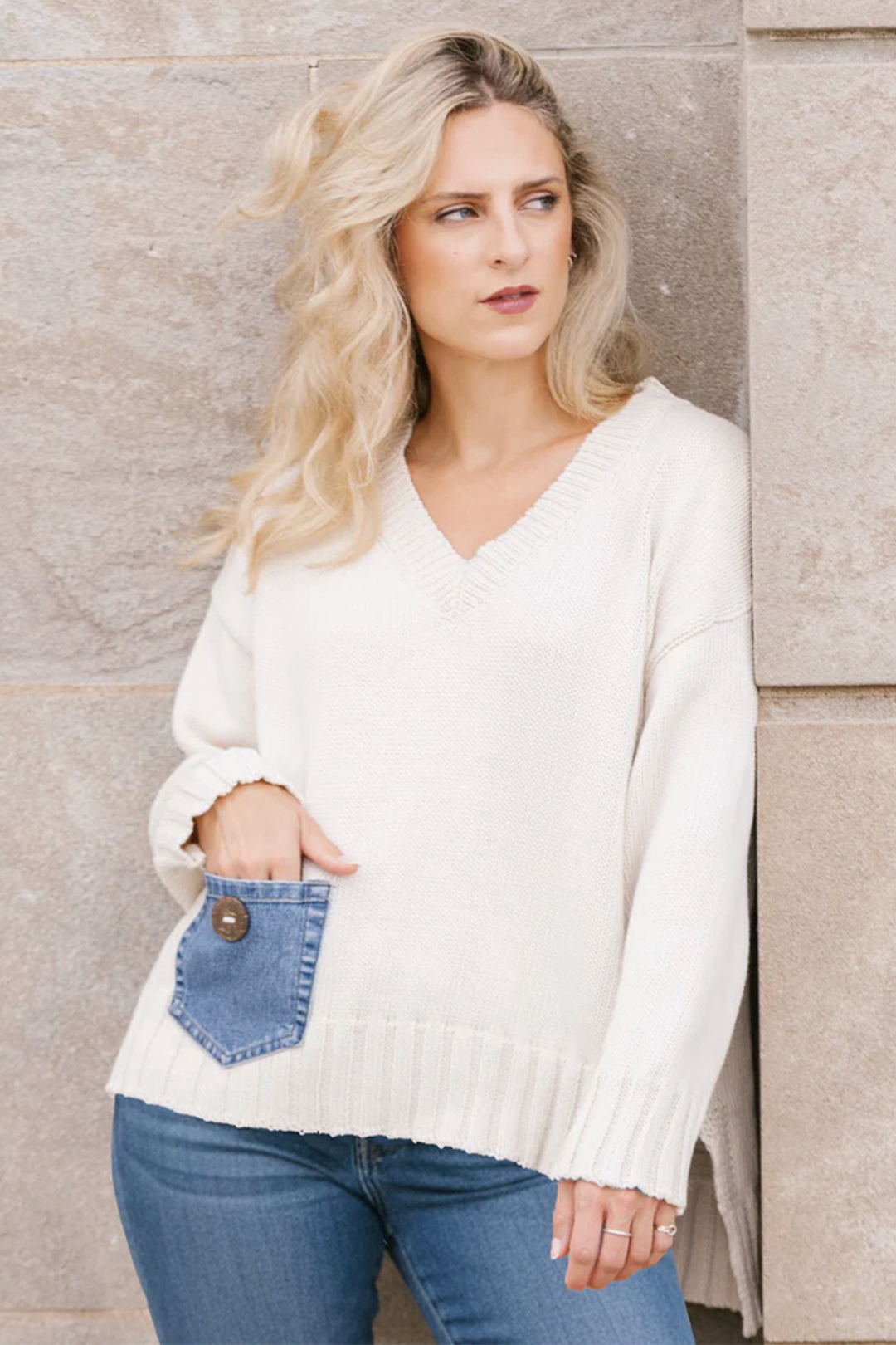 Woman wearing a white sweater with a blue denim patch pocket and jeans, standing against a stone wall.