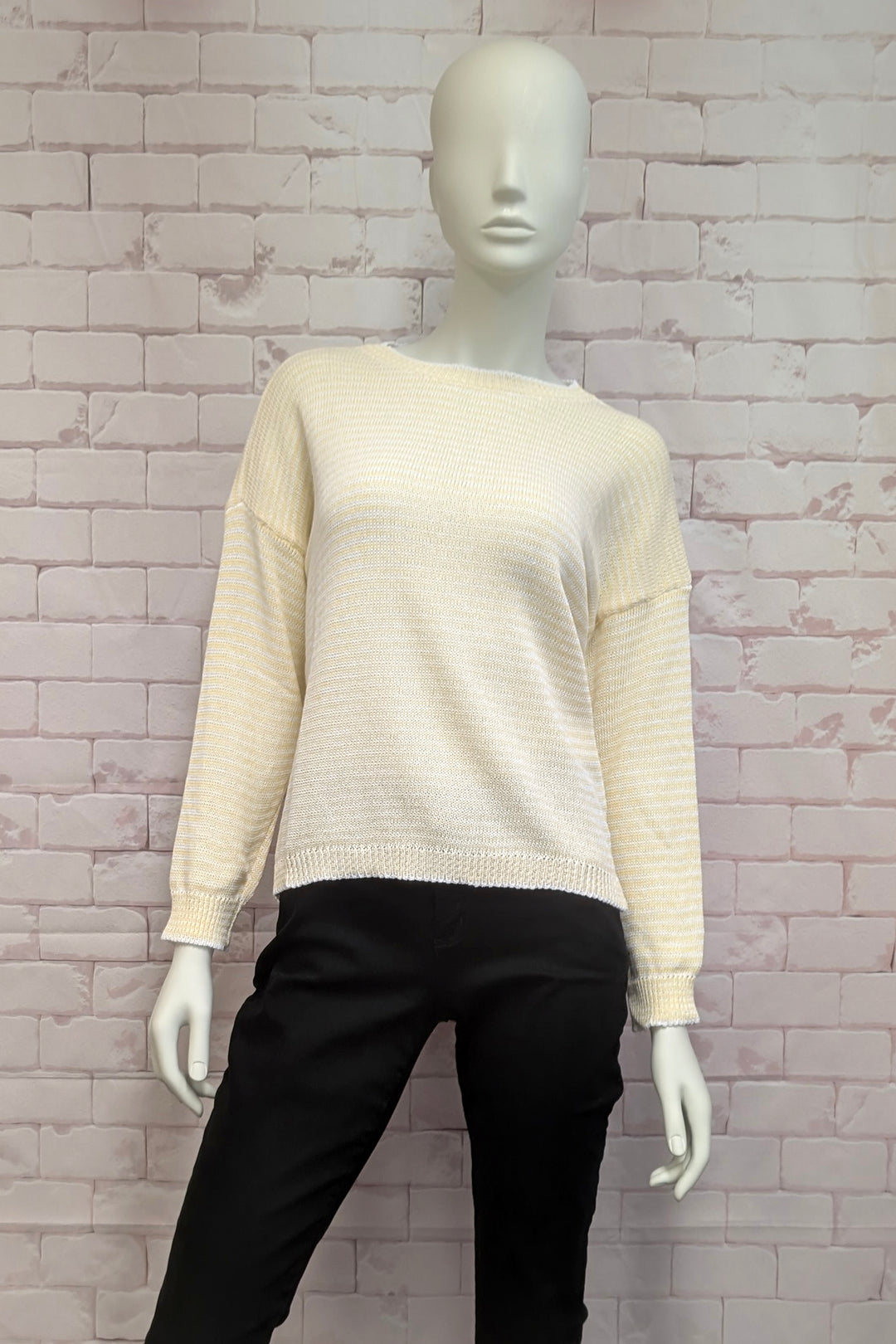 Mannequin wearing a beige light cotton sweater and black pants against a textured wall.