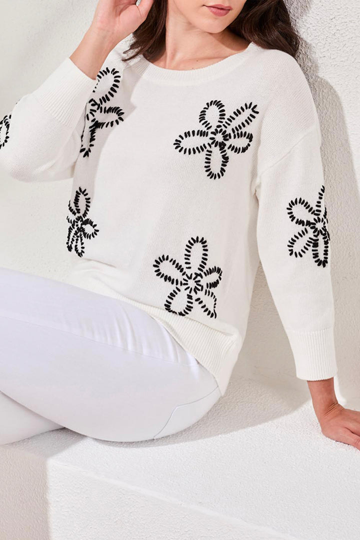 Woman wearing a white light cotton sweater with black floral patterns sitting on a white surface.