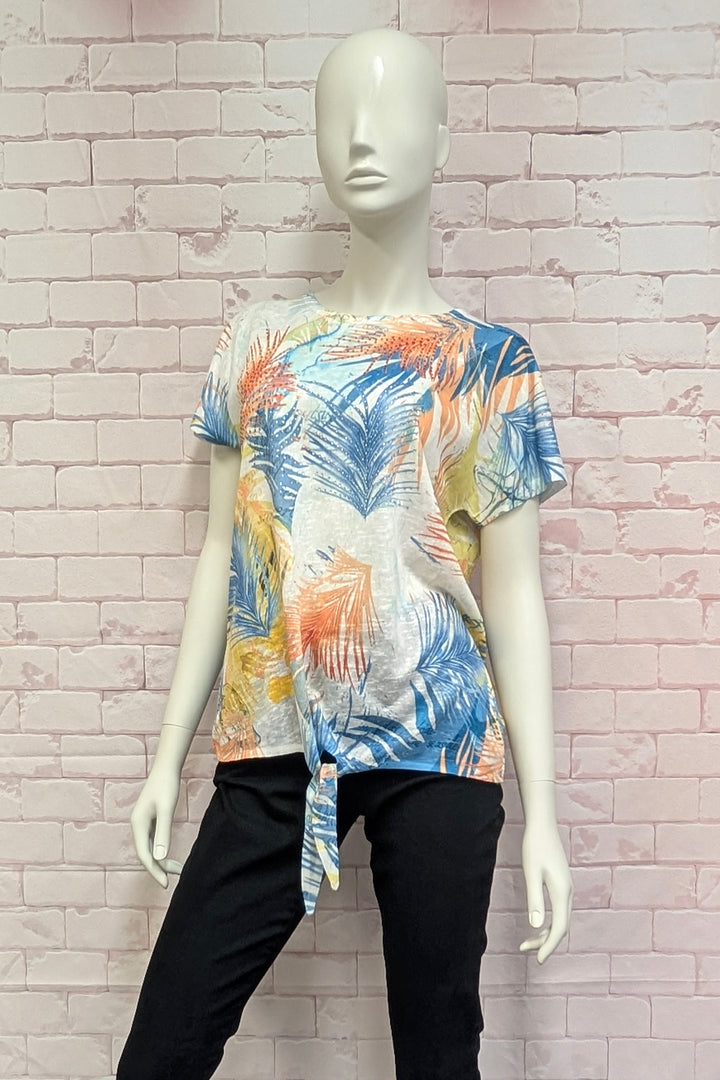 Mannequin wearing a colorful tropical palm print top with tie knot against a light brick wall.
