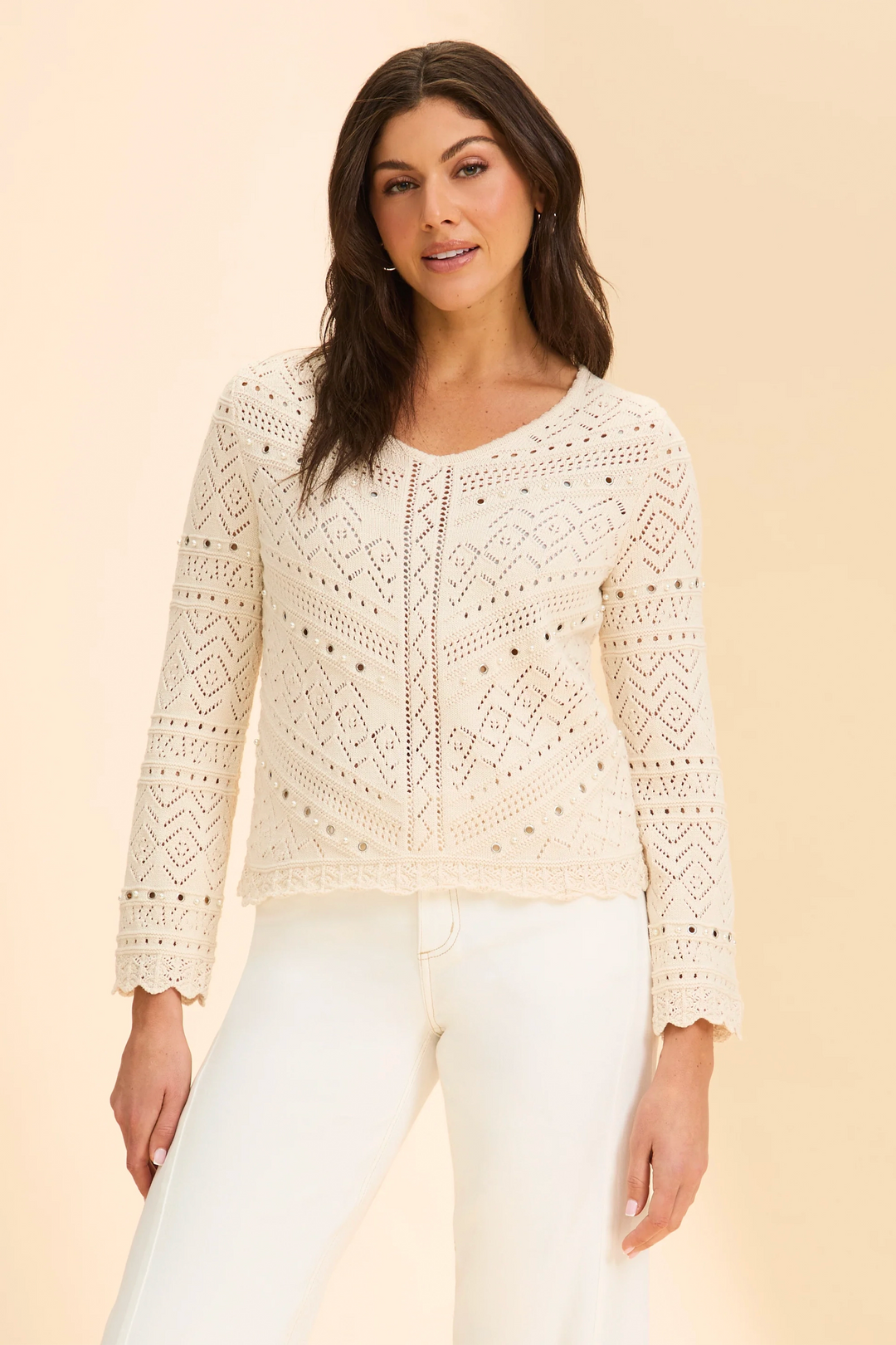 Woman wearing a cream lace top and white pants against a beige background