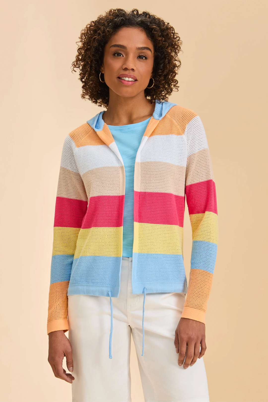 A woman modeling a colorful striped knit zip-up hoodie with a hood, featuring horizontal bands of light blue, yellow, pink, orange, beige, and white, worn over a light blue top and white pants against a solid beige background.