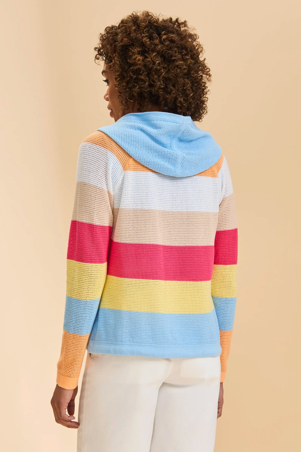 A woman shown from the back wearing a colorful striped knit hoodie with a light blue hood, featuring horizontal stripes of light blue, beige, orange, pink, yellow, and light blue, paired with white pants against a solid beige background.
