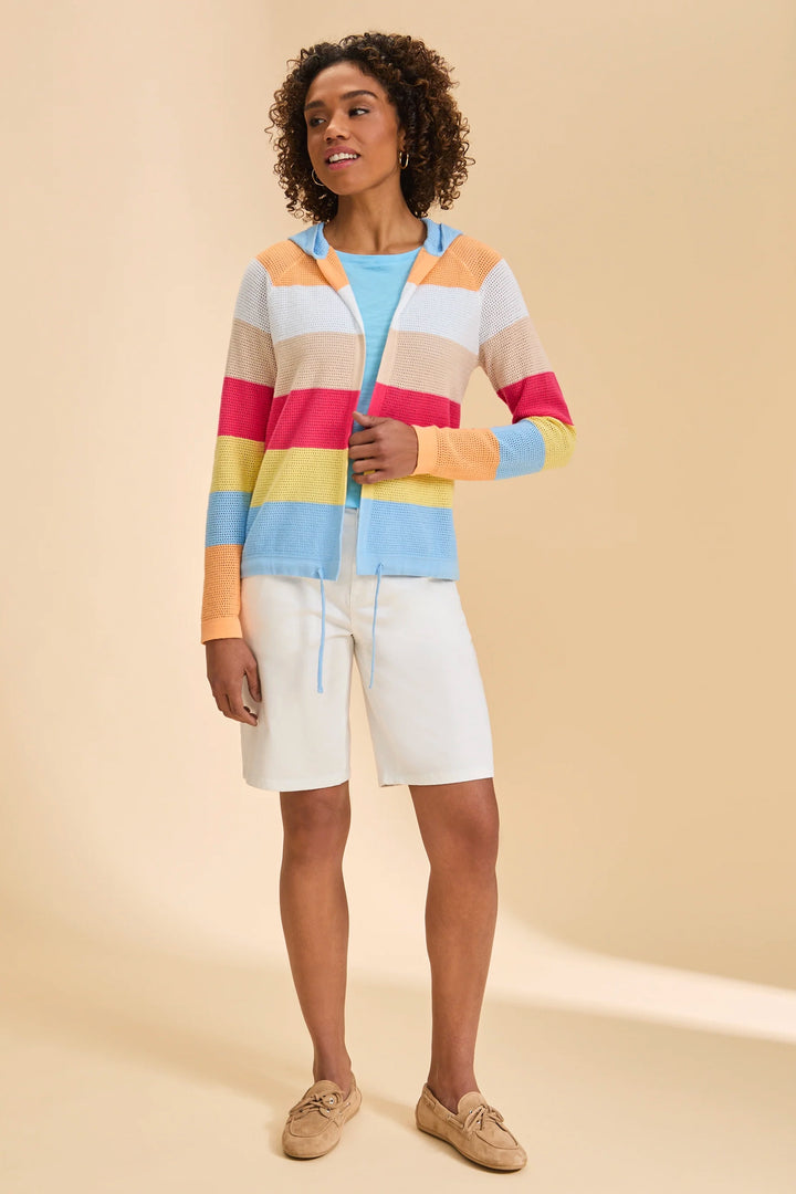 A woman modeling a colorful striped zip-up hoodie with pastel bands of pink, yellow, light blue, orange, and cream, paired with white shorts and tan loafers against a solid beige studio background.
