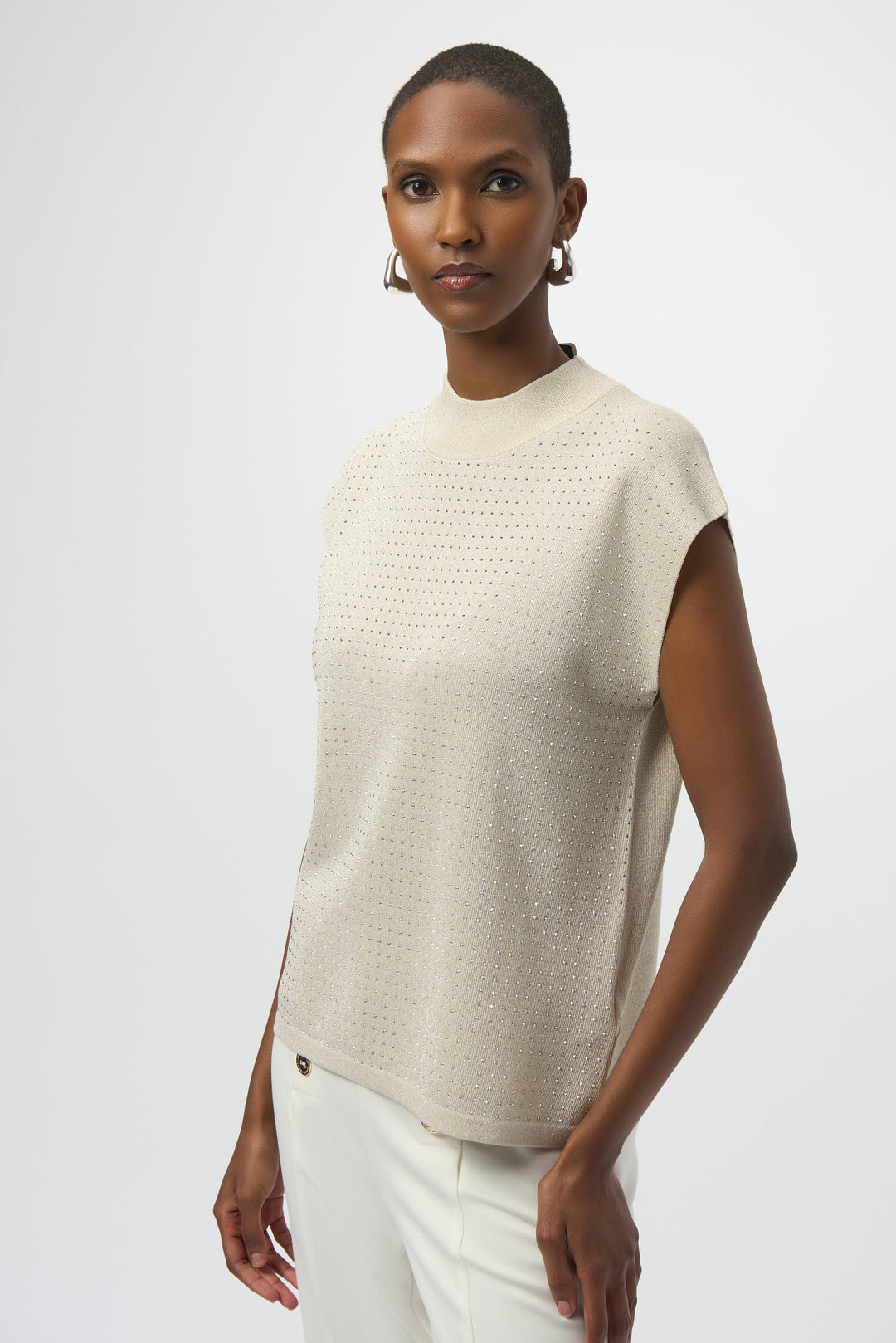 Woman wearing a beige sleeveless top and white pants on a plain background