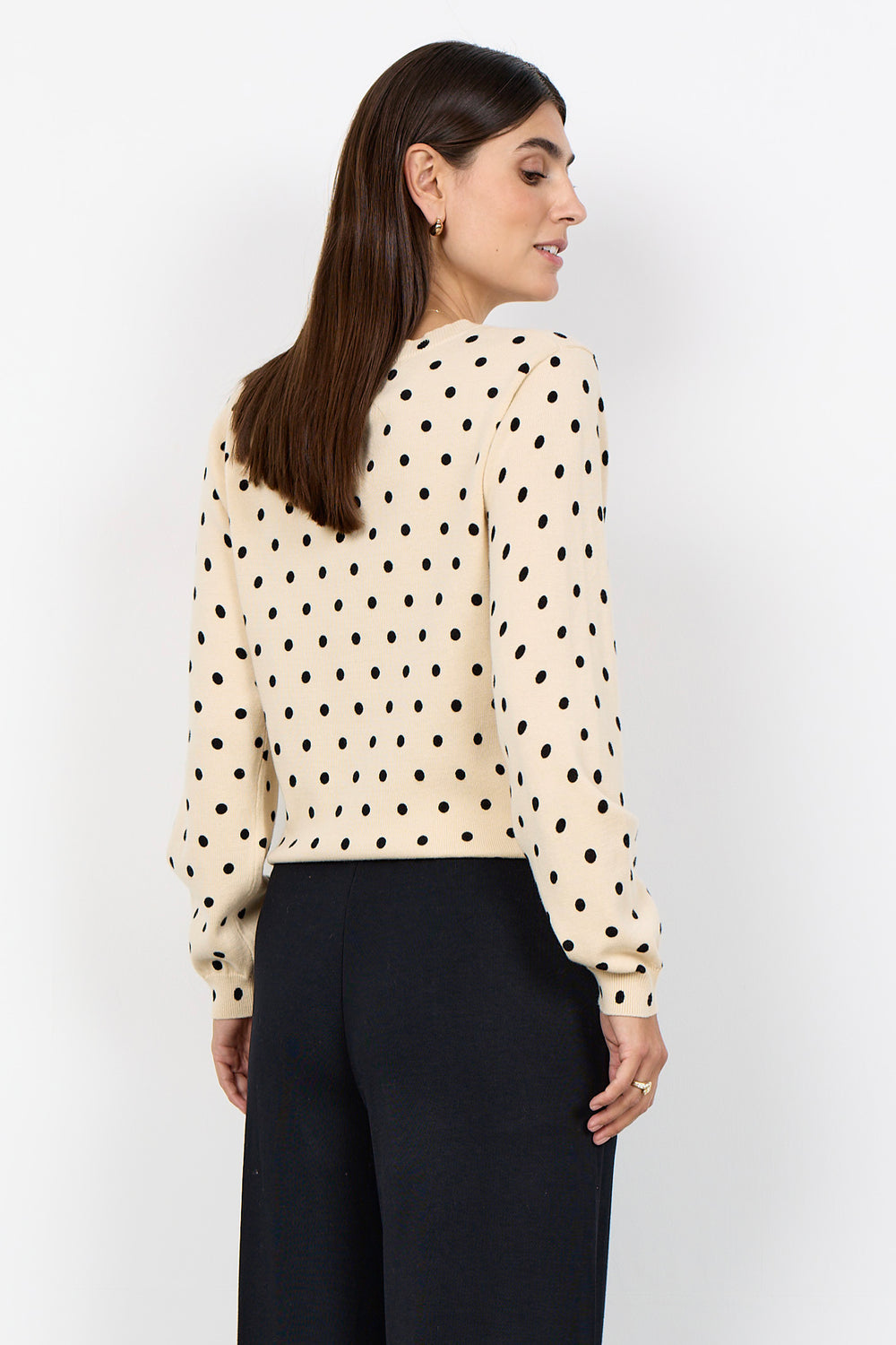 woman wearing a cream cardigan with black polka dots and black pants on a white background. 