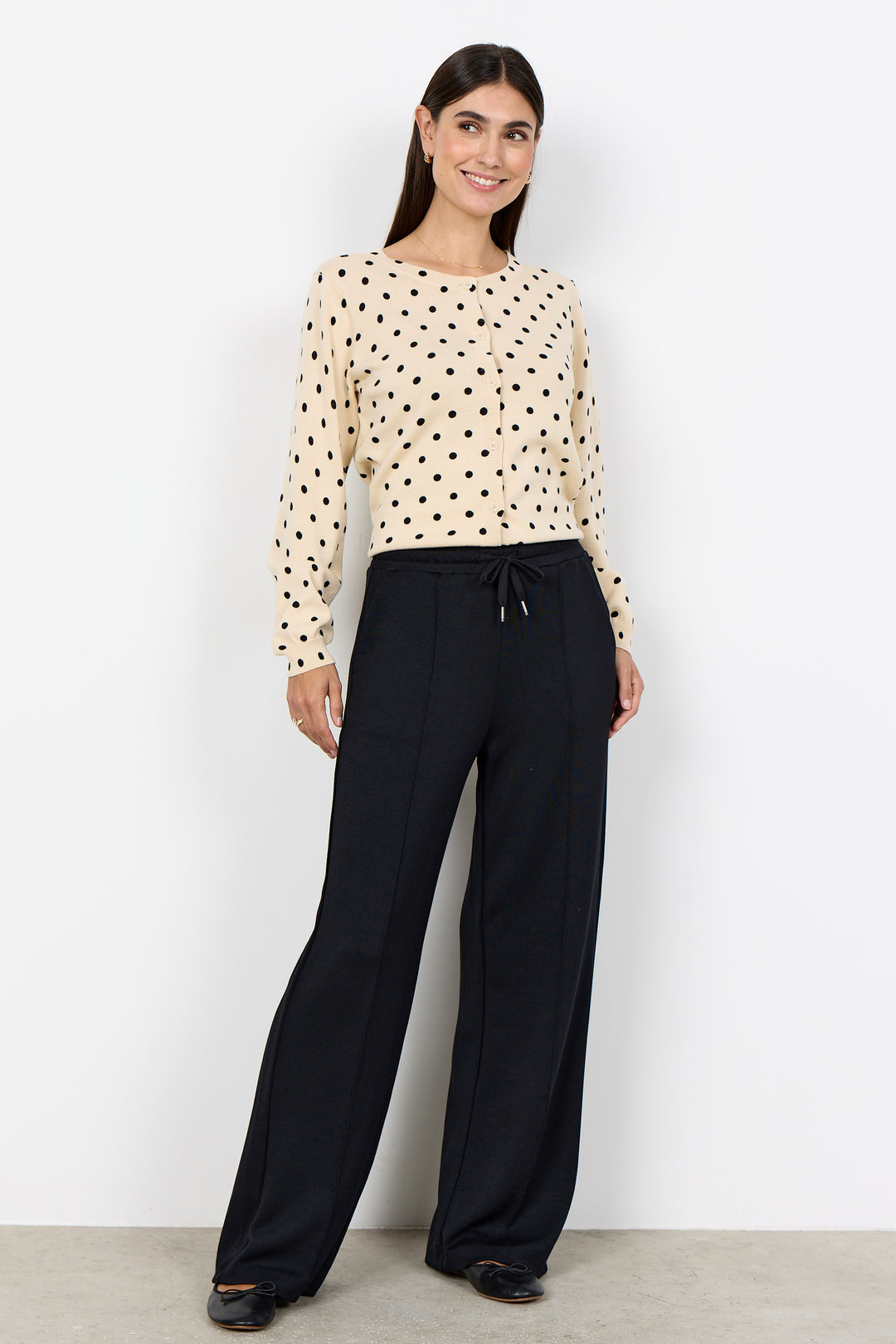 woman wearing a cream cardigan with black polka dots and black pants on a white background. 