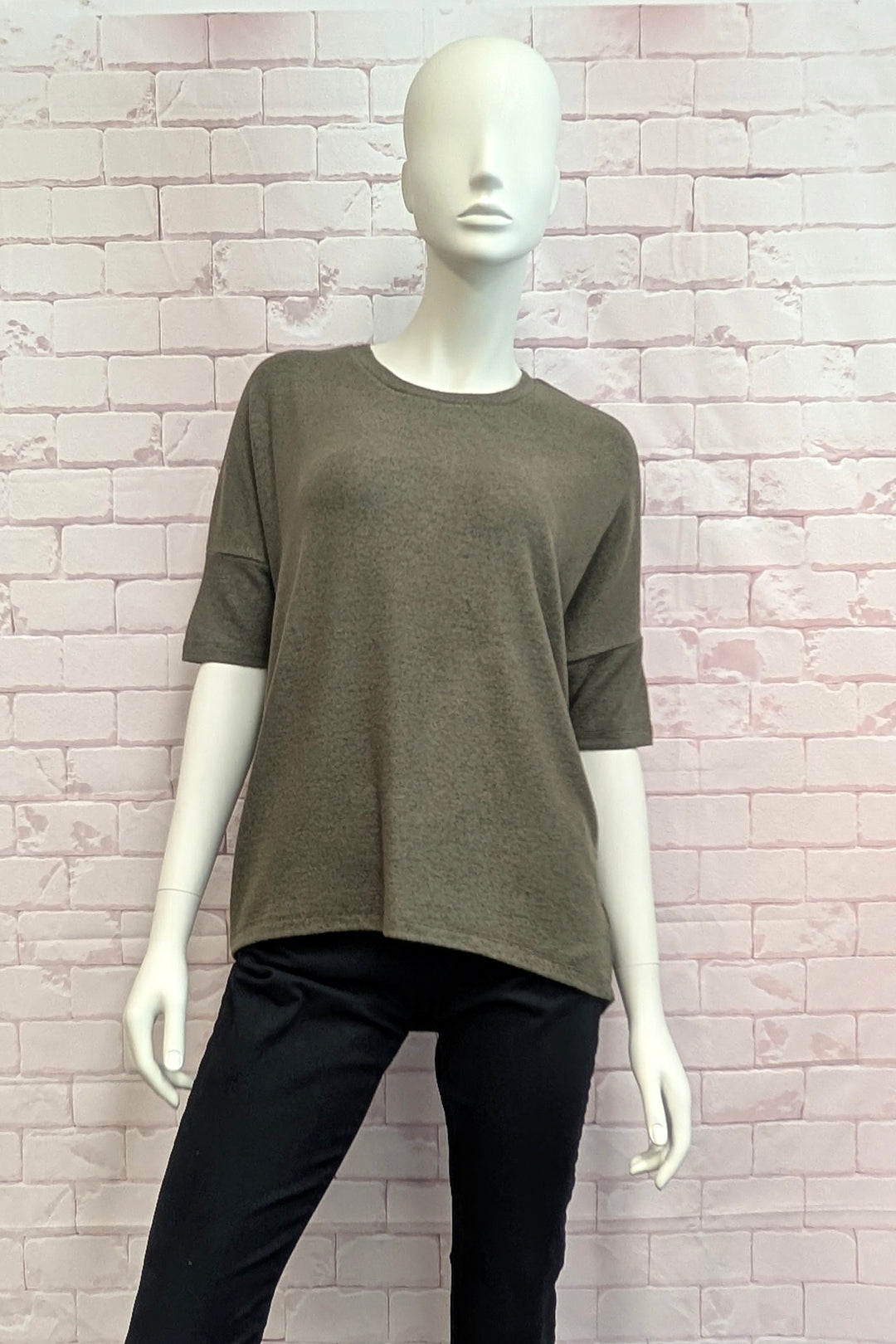 An olive green short-sleeve knit top displayed on a white mannequin, paired with black pants, set against a light pink brick wall background.