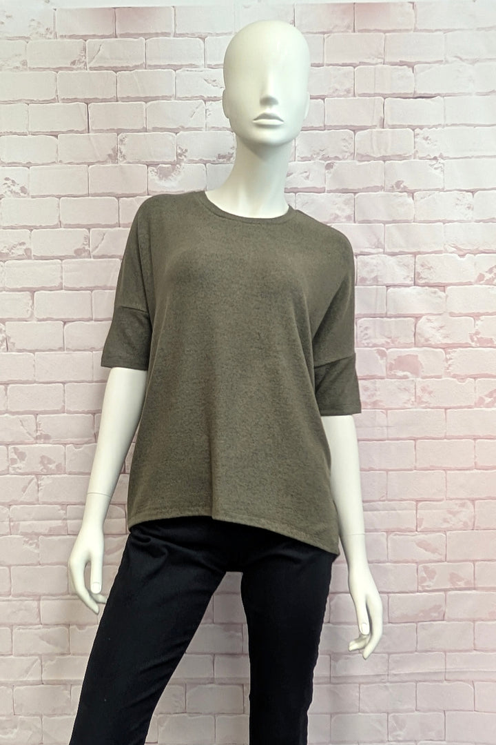 An olive green short-sleeve knit top displayed on a white mannequin, paired with black pants, set against a light pink brick wall background.