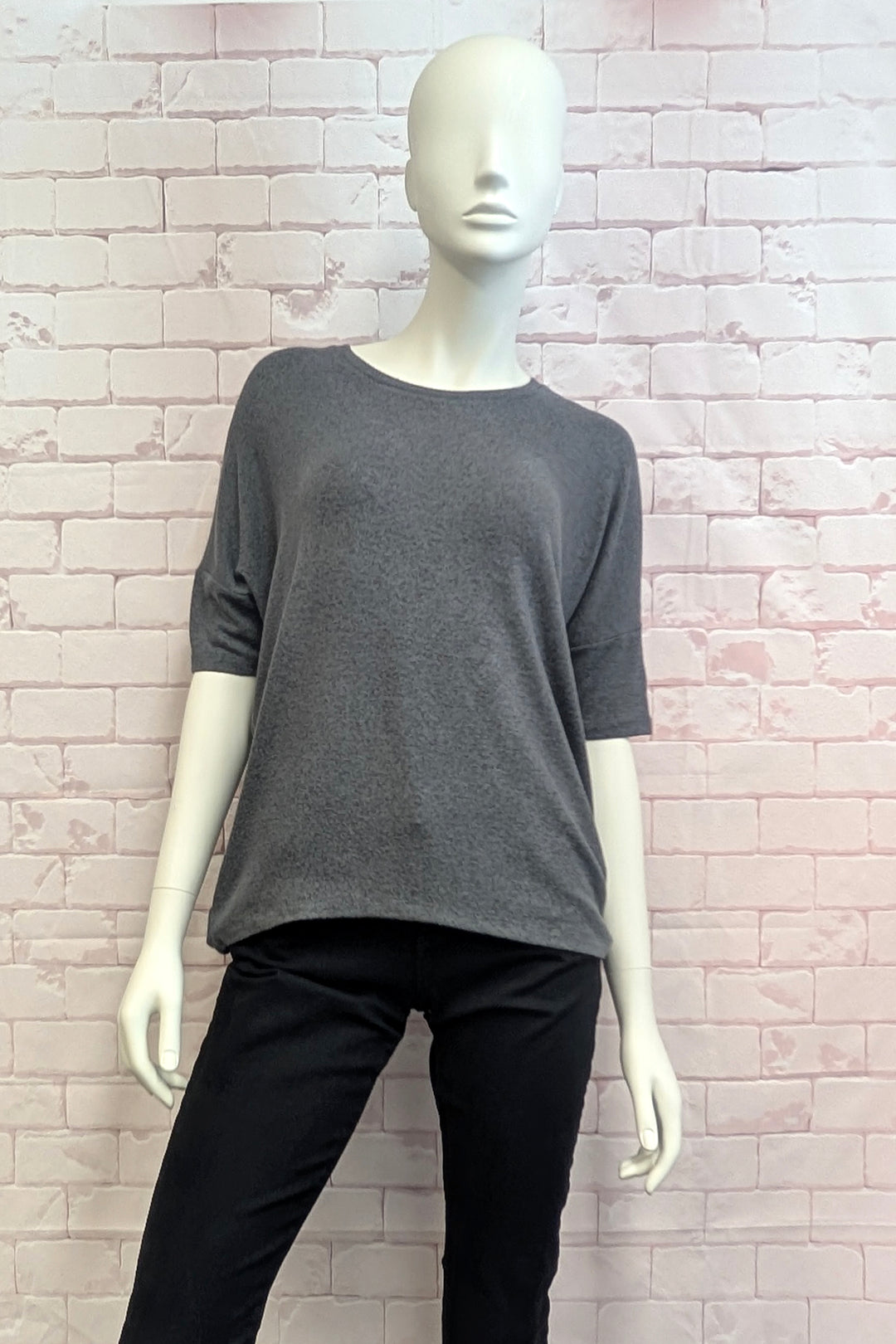A dark gray knit short-sleeve top displayed on a white mannequin, paired with black pants, set against a light pink brick wall background.