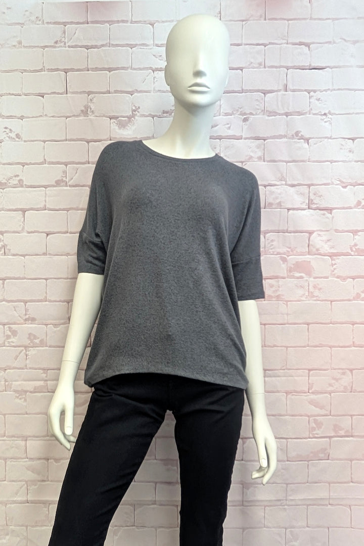 A dark gray knit short-sleeve top displayed on a white mannequin, paired with black pants, set against a light pink brick wall background.