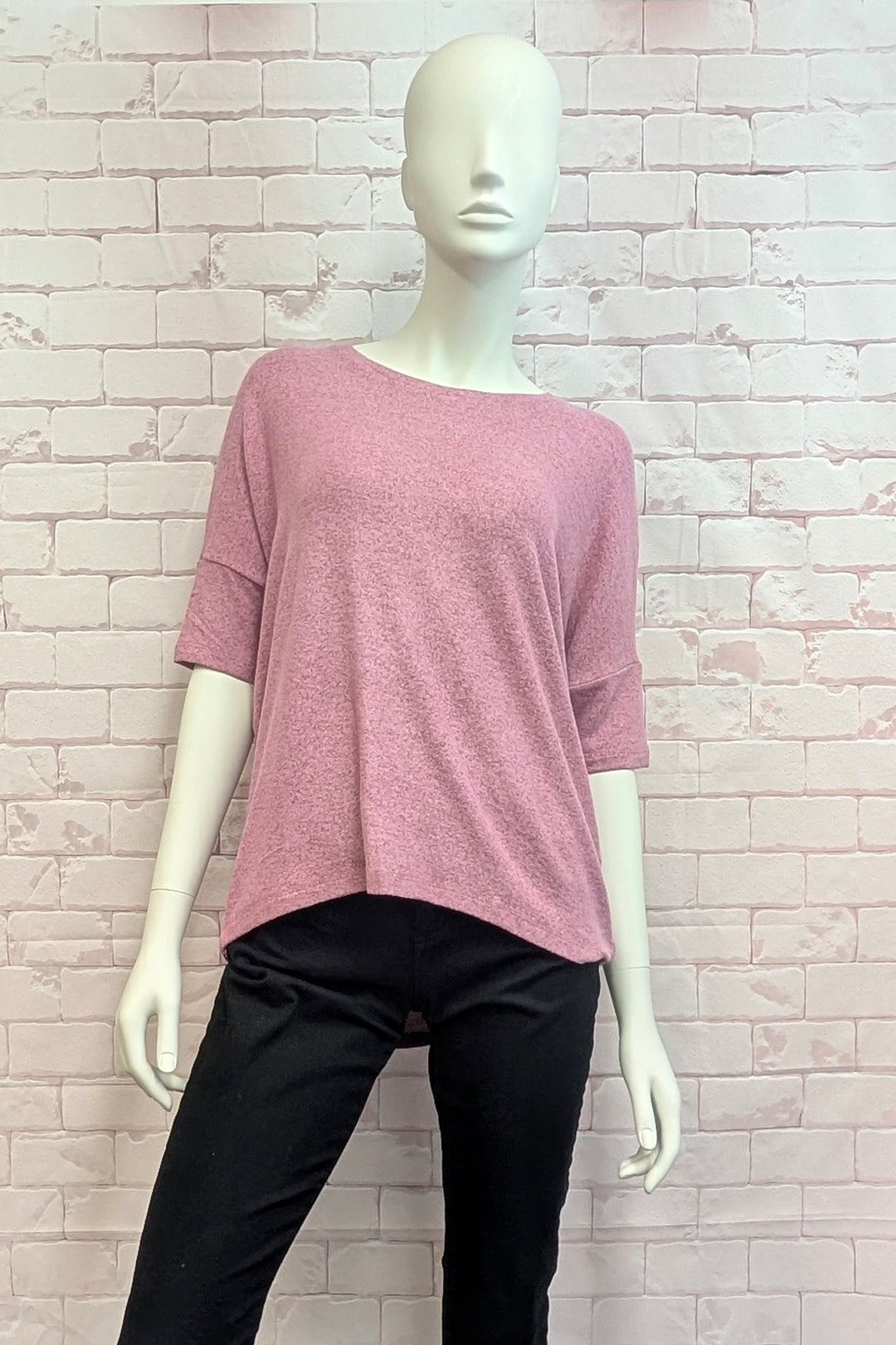 A pink short-sleeve knit top displayed on a white mannequin, paired with black pants, set against a light pink brick wall background.