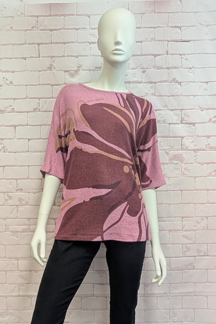 A pink and brown floral print short-sleeve top displayed on a white mannequin, paired with black pants, set against a light brick wall background.
