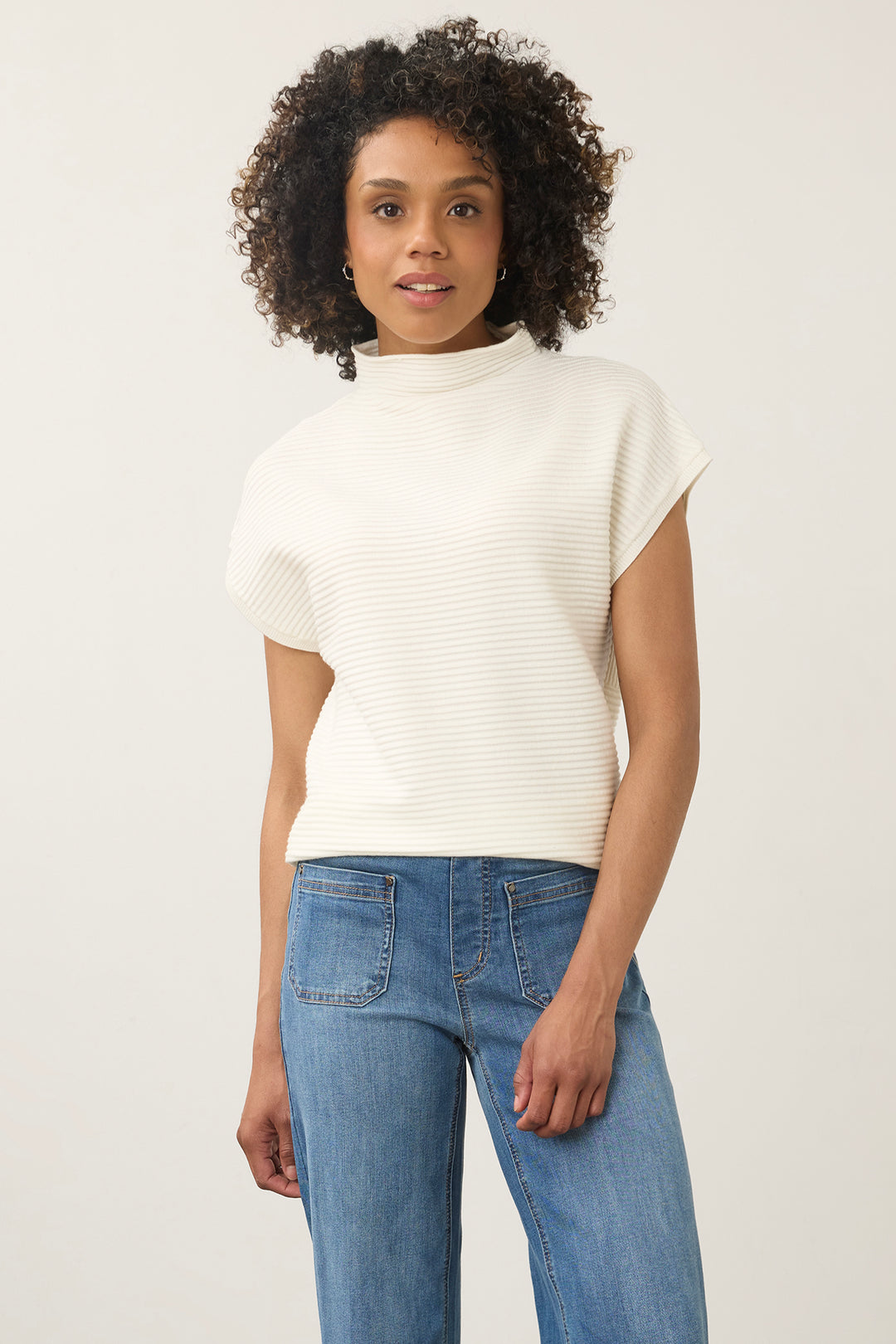 A woman with curly dark hair wearing a cream ribbed short-sleeve mock neck top and blue jeans, standing against a light gray studio background.