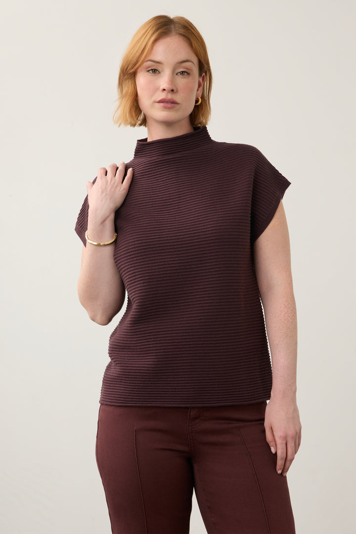 A woman modeling a ribbed, short-sleeve mock neck top in a deep plum-brown color, paired with matching trousers, against a plain light gray studio background.