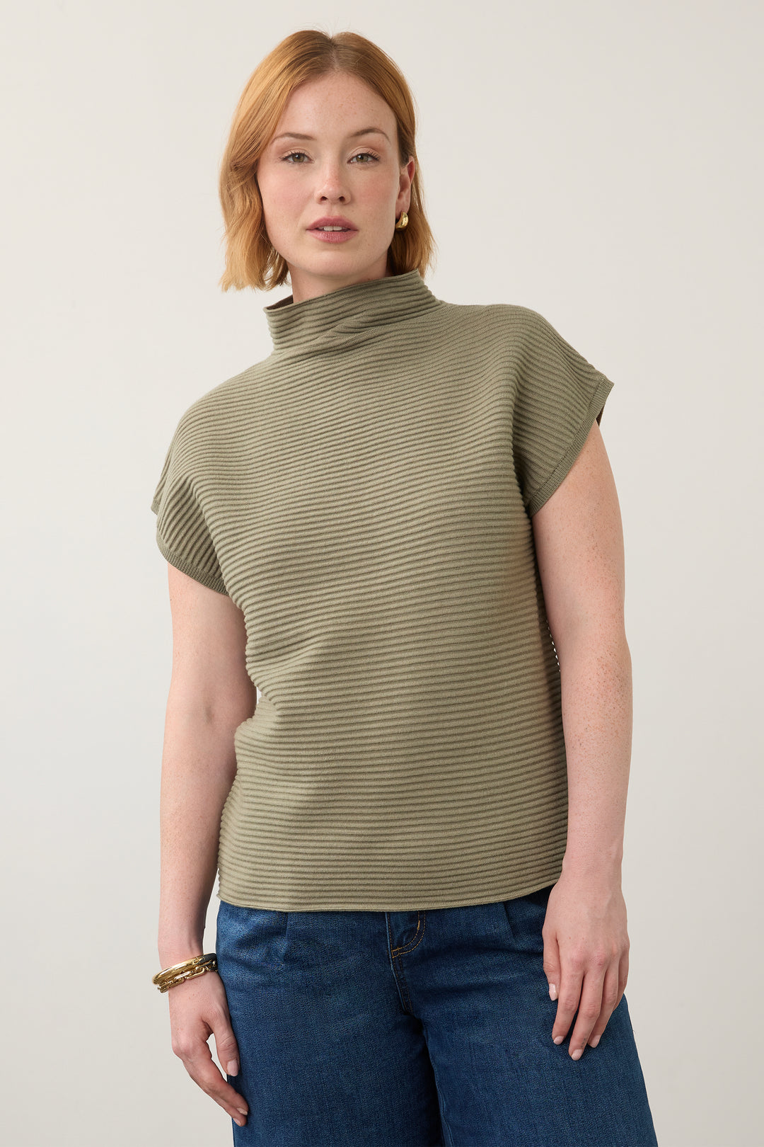 A woman wearing a ribbed olive green mock neck top with short sleeves and blue jeans, standing against a plain light gray background.