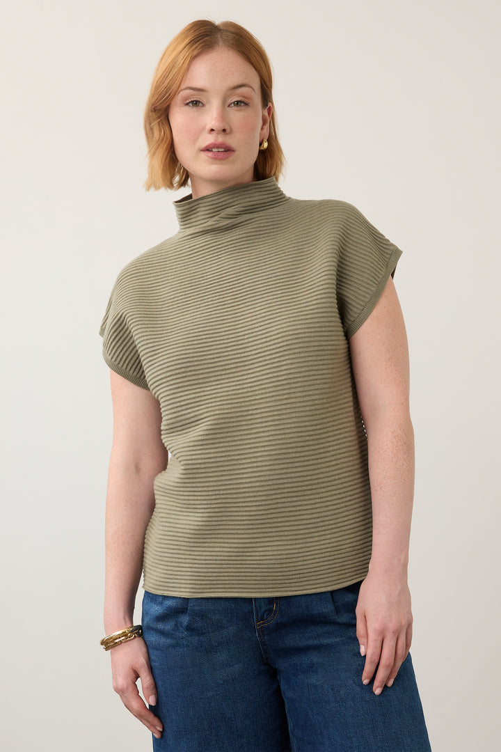 A woman wearing a ribbed olive green mock neck top with short sleeves and blue jeans, standing against a plain light gray background.