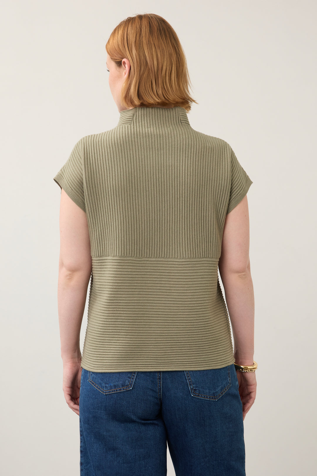 A woman viewed from the back wearing a ribbed short-sleeve mock neck top in olive green, paired with blue jeans, against a plain light background.