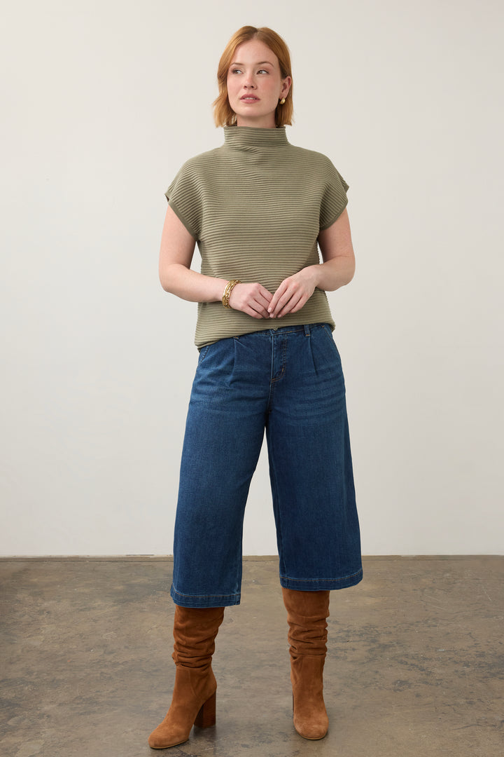 A woman stands in a studio wearing a ribbed olive green short-sleeve mock neck top, blue cropped wide-leg jeans, and brown suede boots.