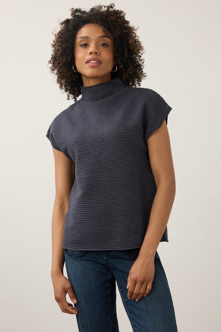 A woman with curly dark hair wearing a dark gray ribbed mock neck top with cap sleeves and blue jeans, standing against a plain light gray studio background.