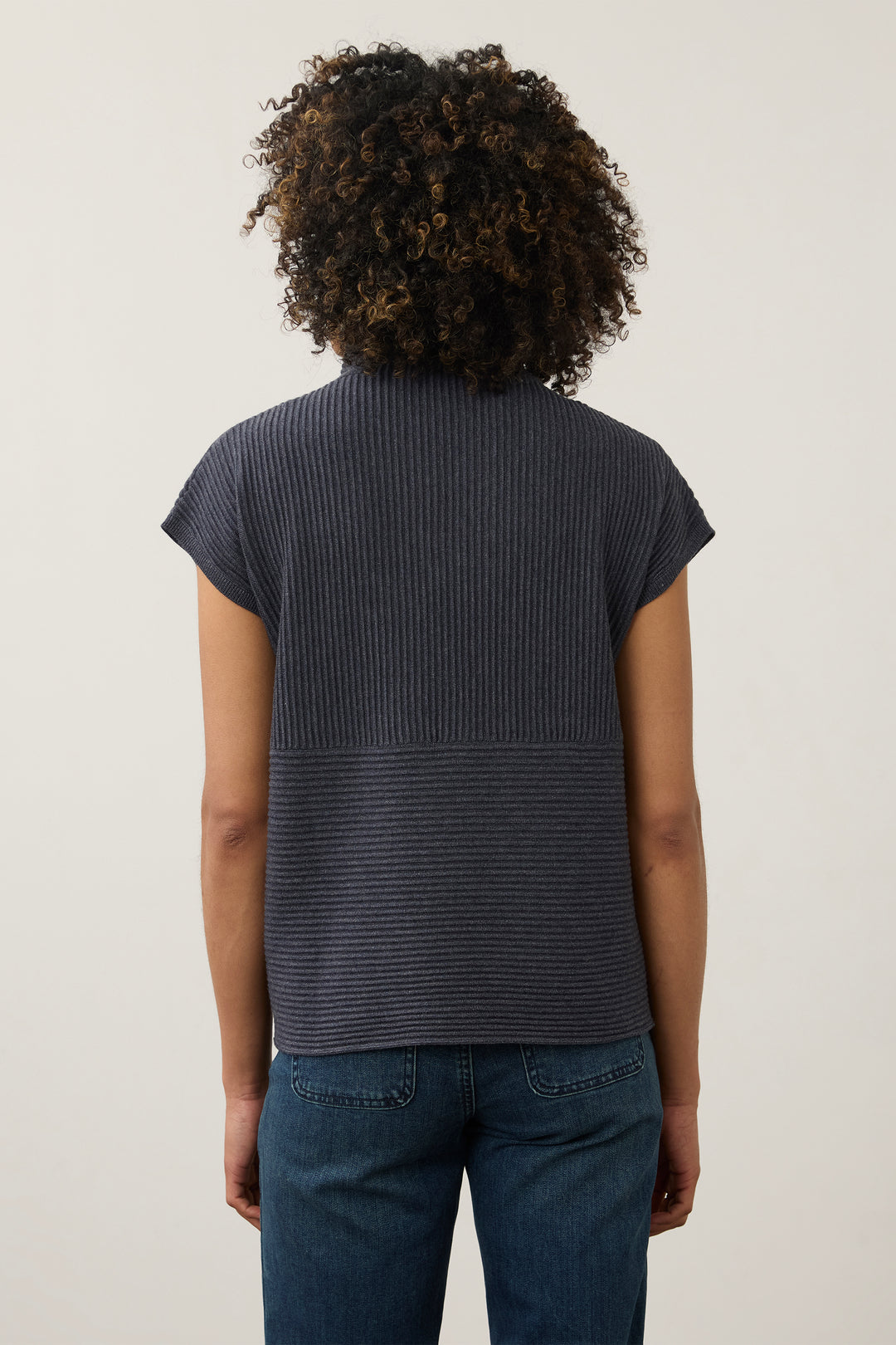 Back view of a person with curly dark hair wearing a dark navy ribbed short-sleeve top and blue jeans against a plain light background.
