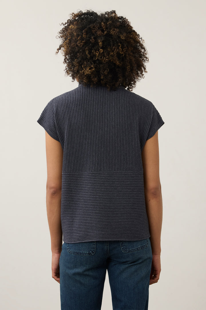 Back view of a person with curly dark hair wearing a dark navy ribbed short-sleeve top and blue jeans against a plain light background.