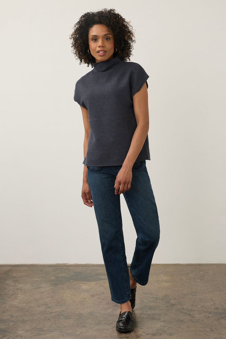 A woman modeling a dark navy ribbed short-sleeve mock neck top with dark blue jeans and black loafers, standing against a plain off-white wall.
