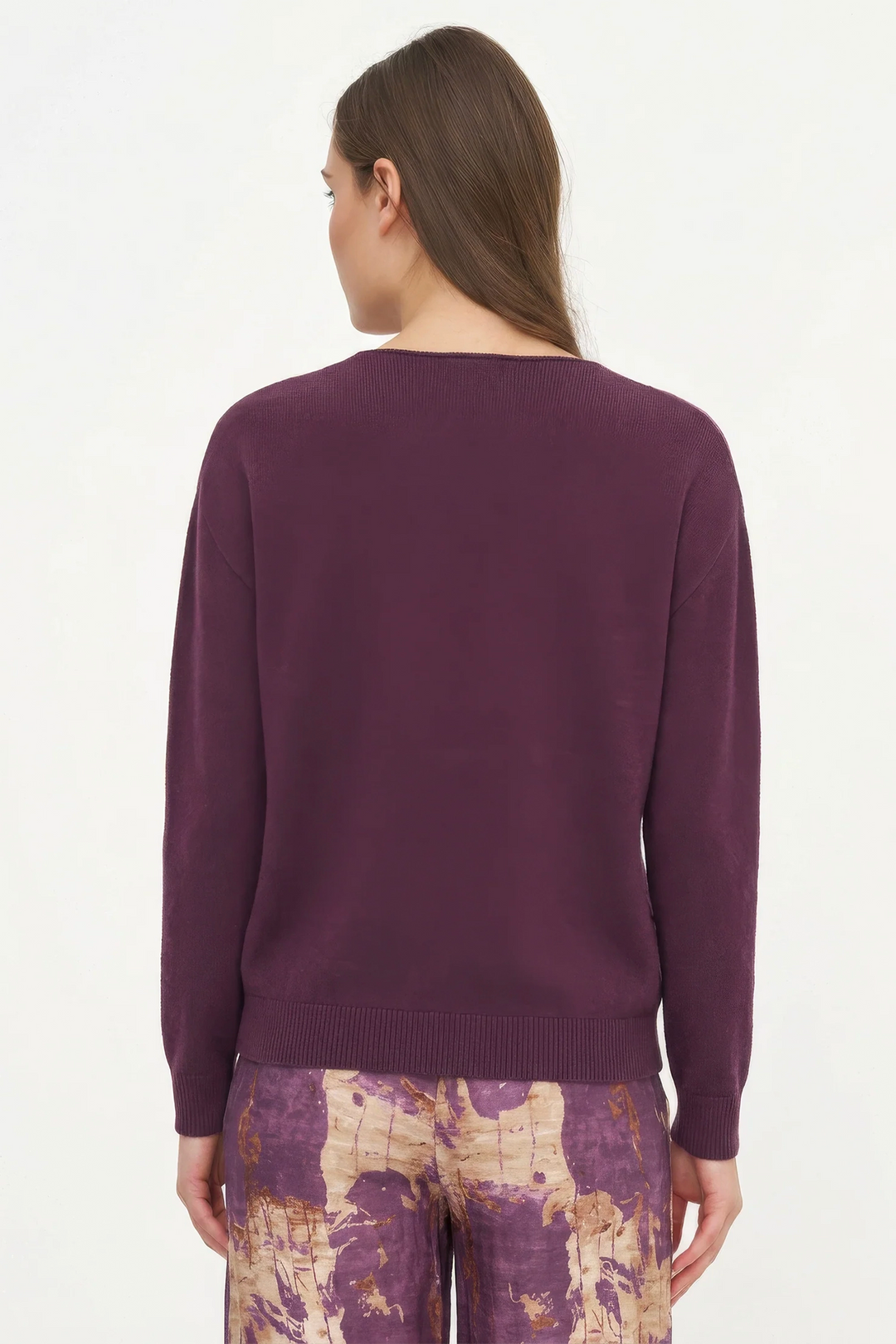 A woman shown from the back wearing a solid deep purple long-sleeve knit sweater and patterned purple and beige pants, against a plain white studio background.