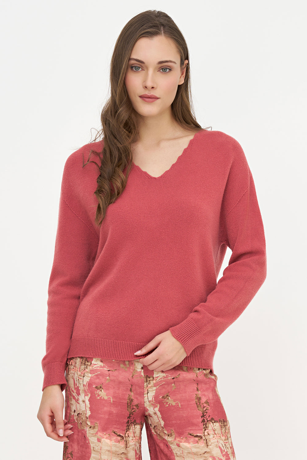 A woman modeling a dusty rose pink V-neck knit sweater with long sleeves, paired with patterned trousers, against a plain white studio background.