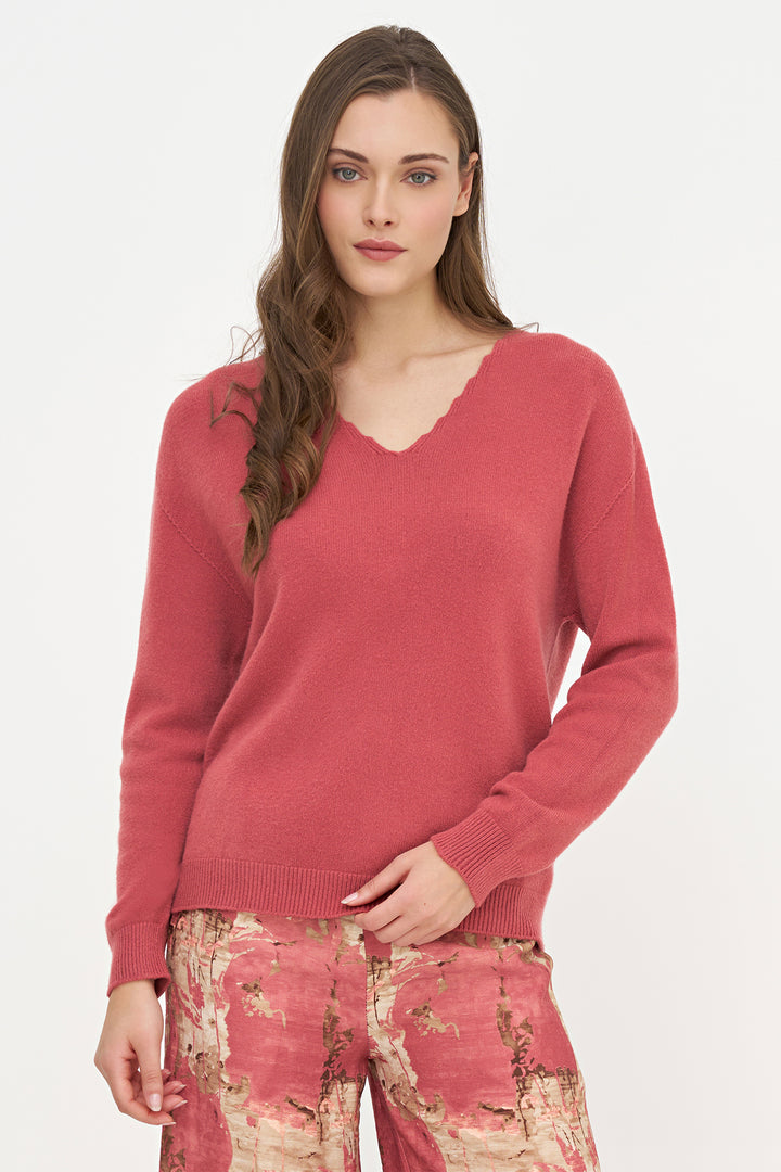 A woman modeling a dusty rose pink V-neck knit sweater with long sleeves, paired with patterned trousers, against a plain white studio background.