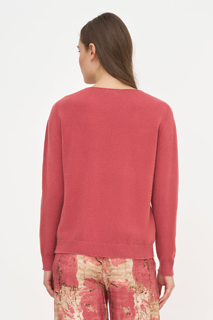 A woman shown from the back wearing a dusty rose pink knit sweater and patterned trousers against a plain white studio background.